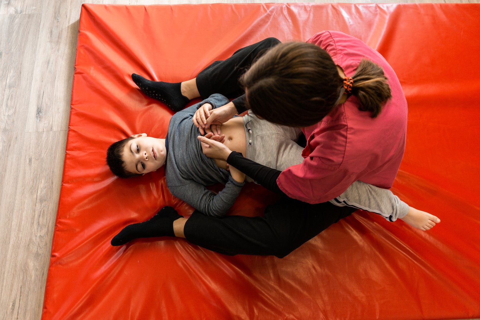 a boy with multiple intellectual and physical disabilities and a female physiotherapist perform exercises on a red mat, she is wearing a uniform and a protection mask. cerebral palsy, reduced mobility