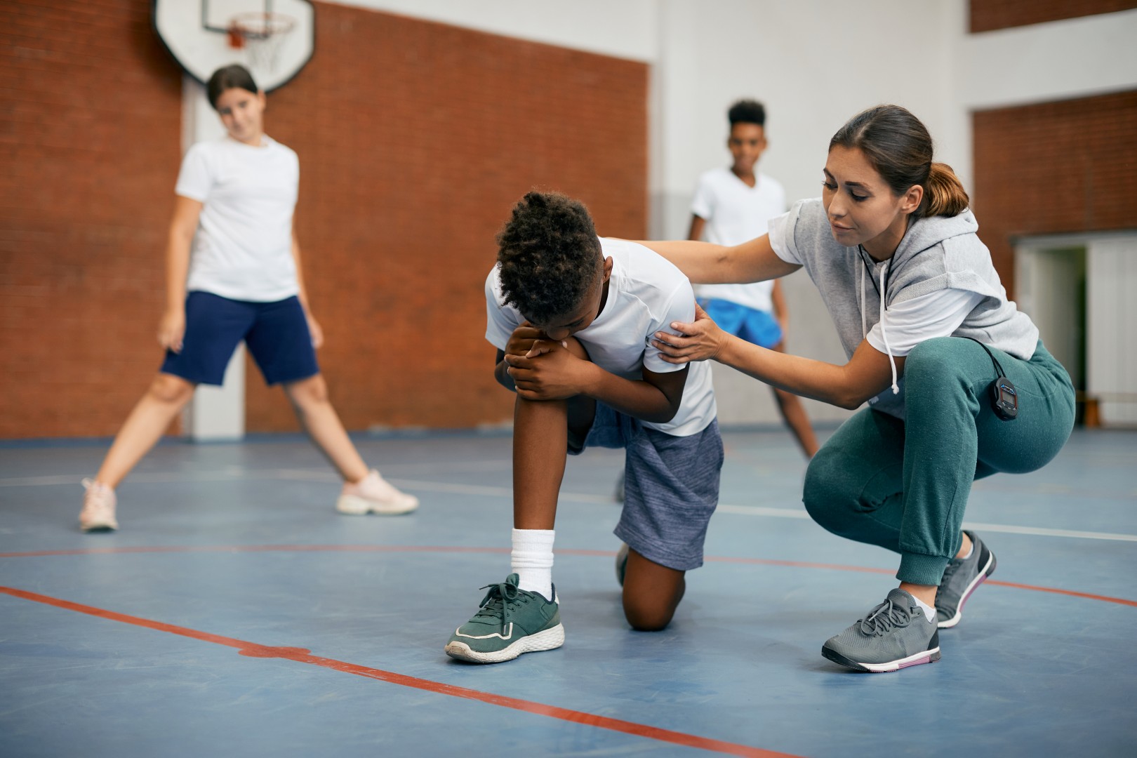 African American schoolboy holding his leg in pain while PE teacher is consoling him during a class at school gym.
